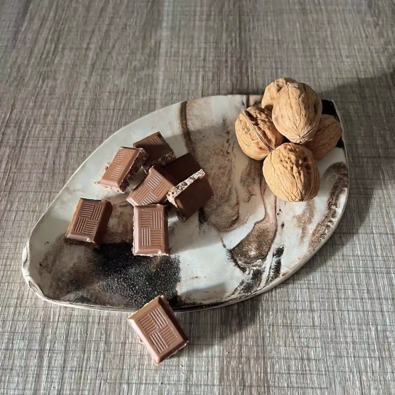 Des carrés de chocolat et des noix disposés dans 1 coupelle en forme de feuille en faience stracciatella